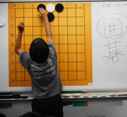 ホワイトボードに貼った囲碁盤に碁石を打つ子どもの写真