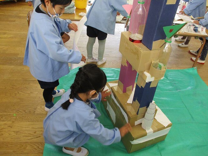 様々な素材で作ったお城に画用紙を貼っている子どもの写真