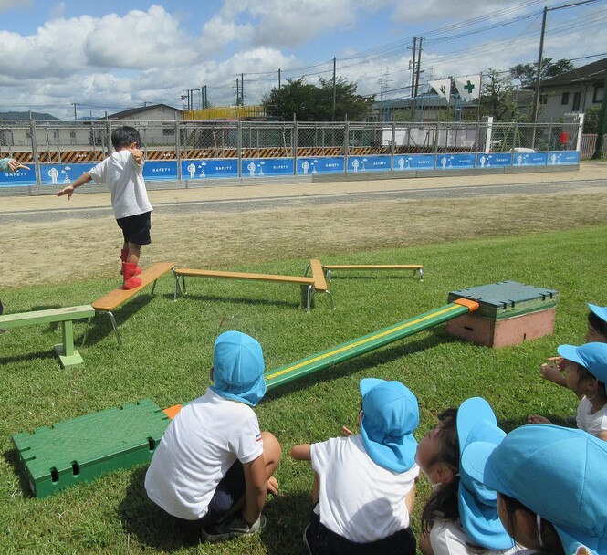 バッタになって、草に見立てた平均台を渡る子どもとそれを見ている子どもの写真